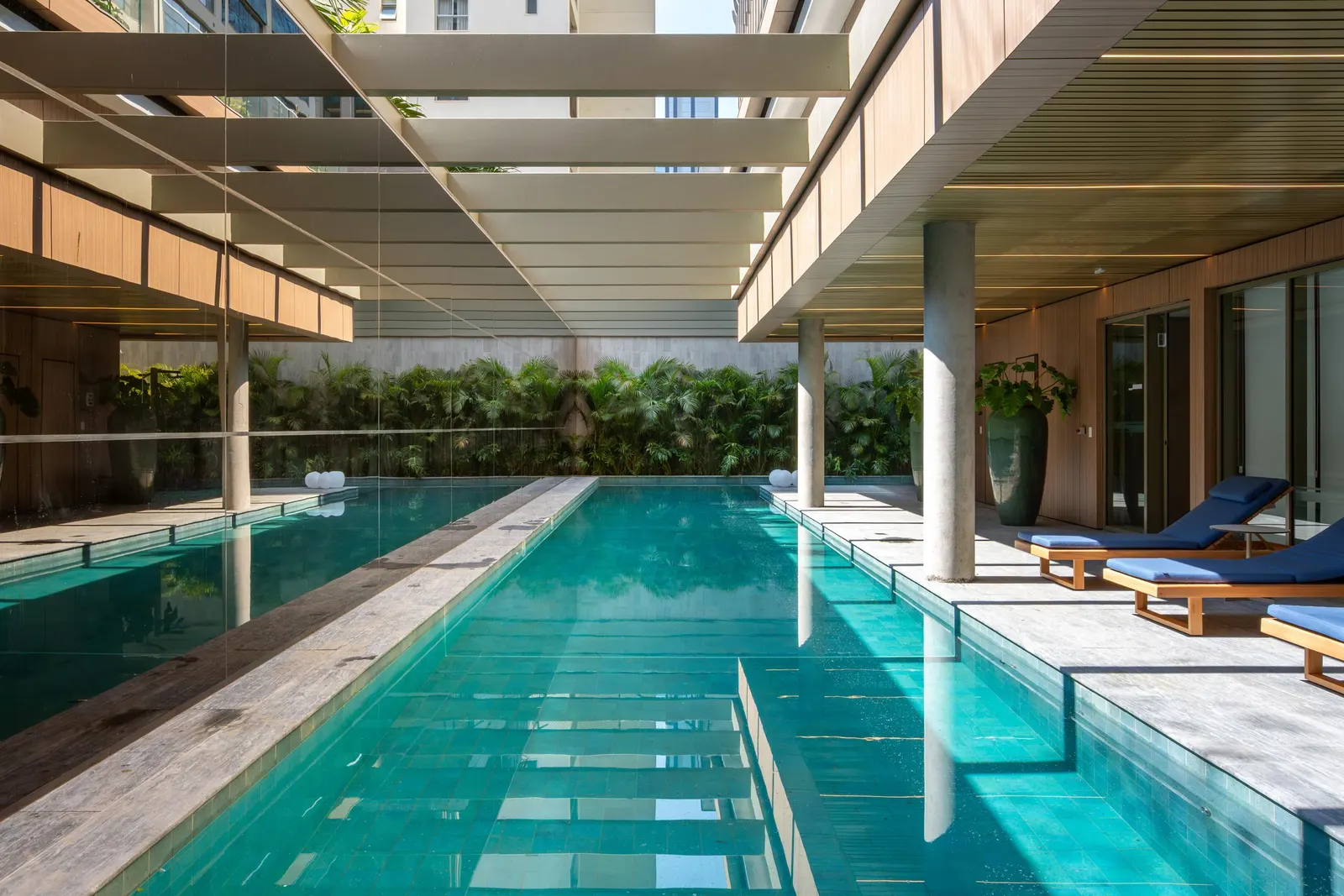 A raia da piscina vista de uma ponta, com as espreguiçadeiras azuis de um lado e o jardim no fundo.