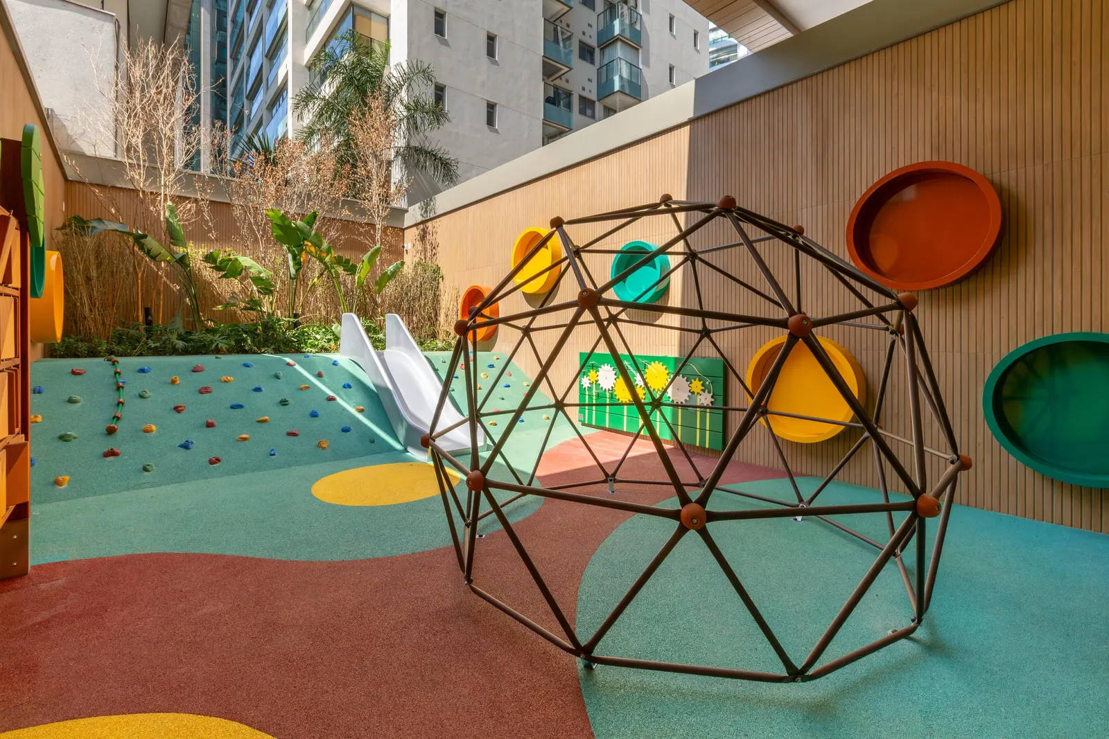 O playground ao ar livre: um domo de escalar, círculos coloridos na parede de madeira e o piso emborrachado em verde e vermelho.