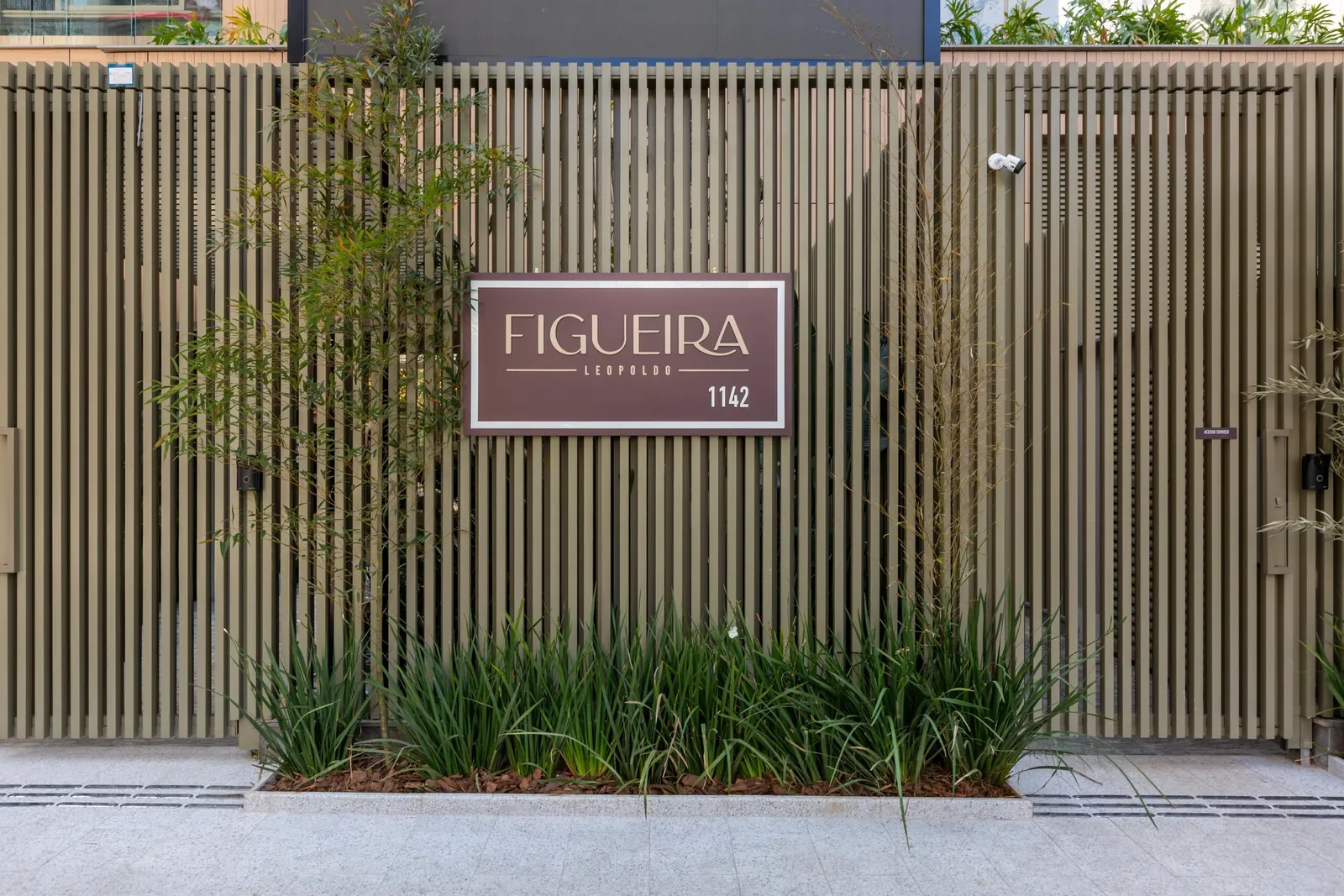 O muro de ripas da portaria, com a placa do Figueira Leopoldo e o número 1142, e um canteiro de plantas na calçada.