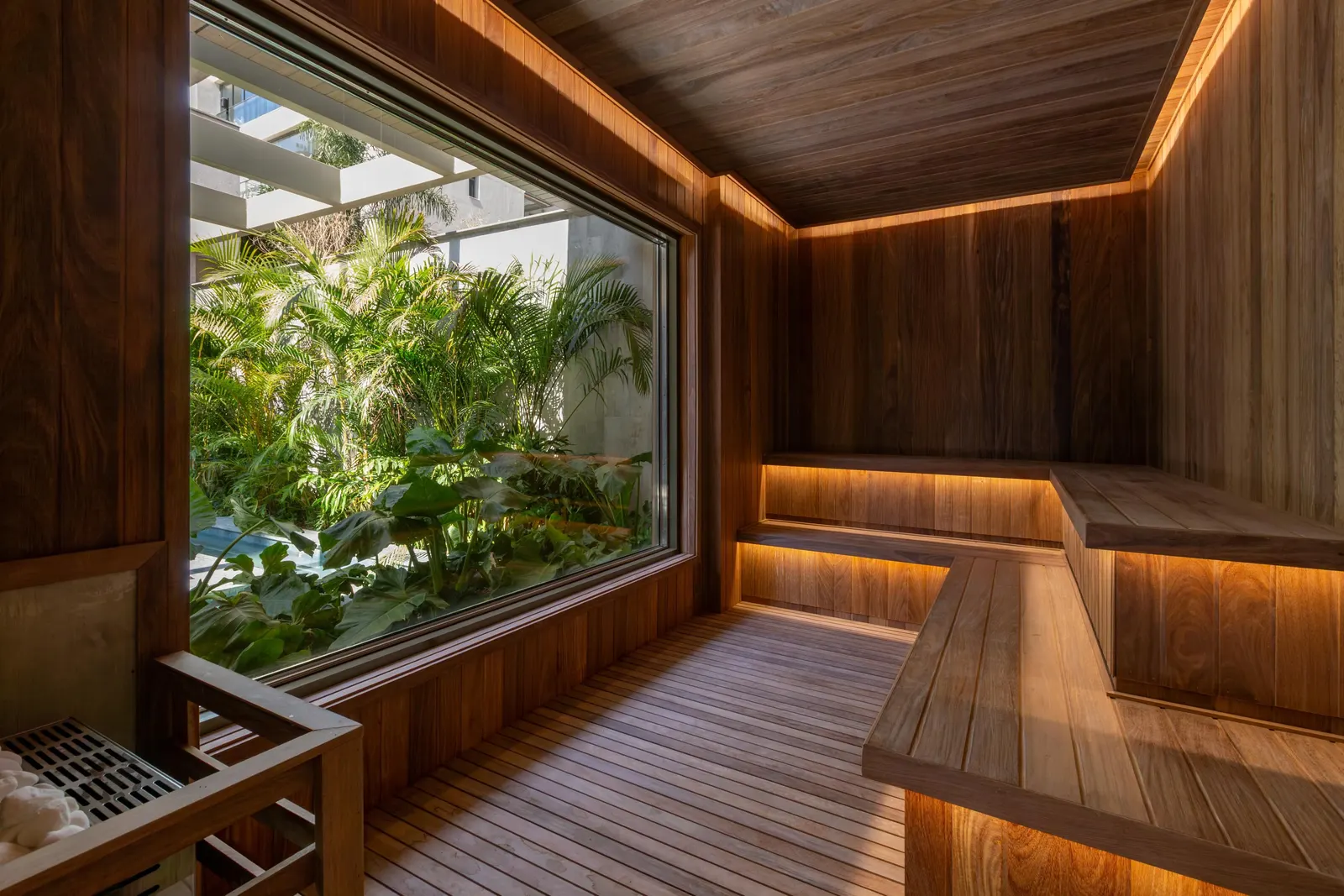A sauna revestida de madeira, com os bancos iluminados por baixo e uma janela grande para o jardim.