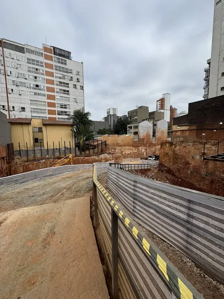 O terreno escavado, com a contenção de concreto à direita e os prédios vizinhos ao fundo, sob o céu nublado.