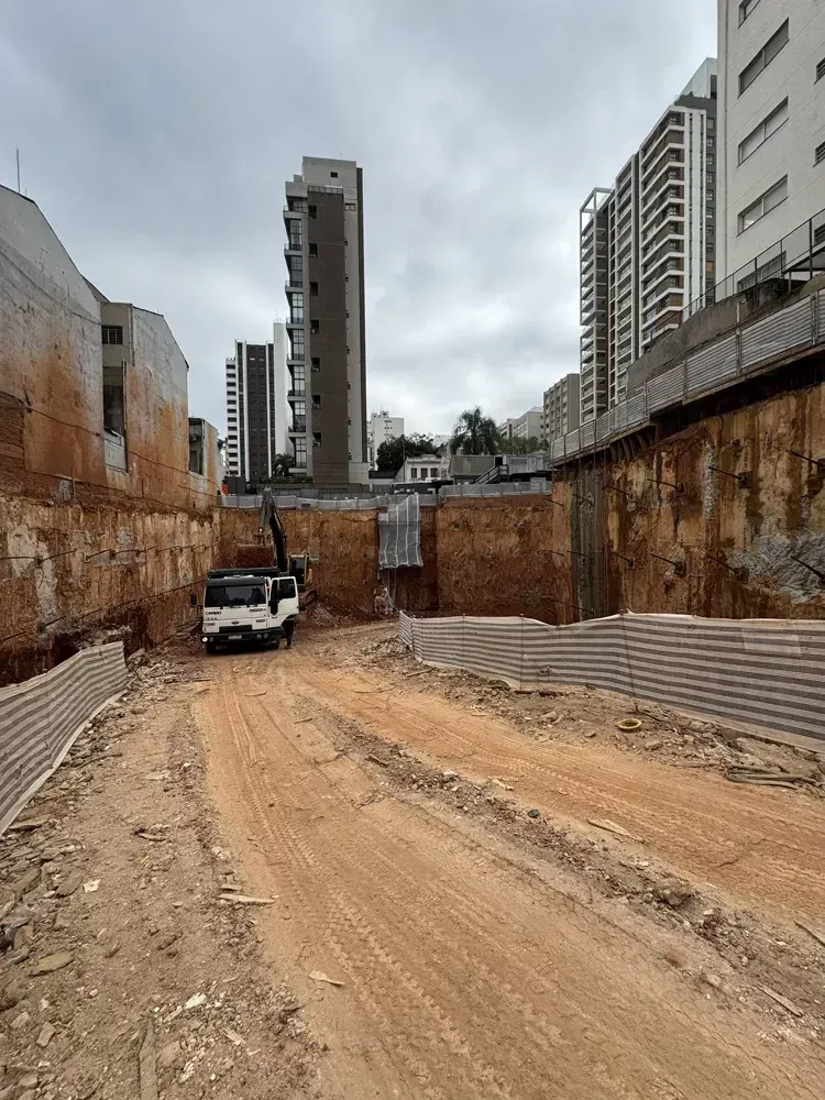 O fundo da escavação com uma caminhonete branca e as paredes de terra e concreto, com os prédios ao fundo.
