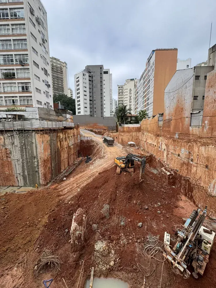 A escavação vista do alto: a terra vermelha, uma escavadeira e uma perfuratriz, com os prédios vizinhos em volta.