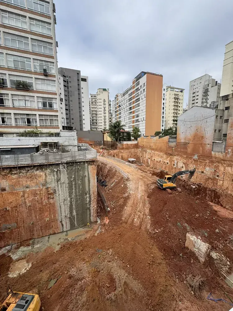A escavação vista do alto, com as máquinas no fundo e a parede de contenção ao lado dos prédios.