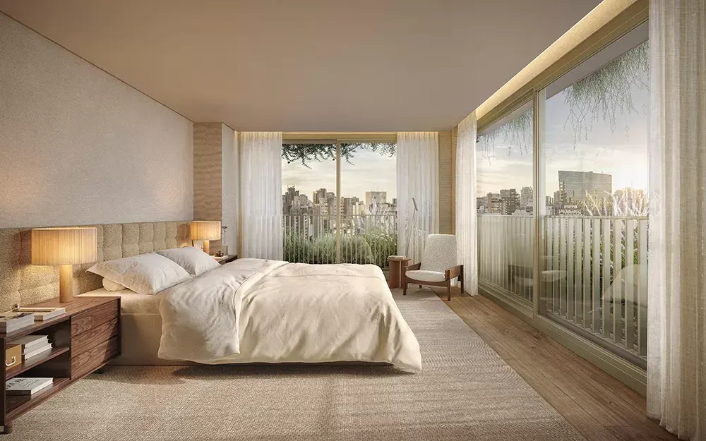 Uma suíte com a cama de frente para o vidro do piso ao teto, cortinas claras e os prédios do bairro lá fora.