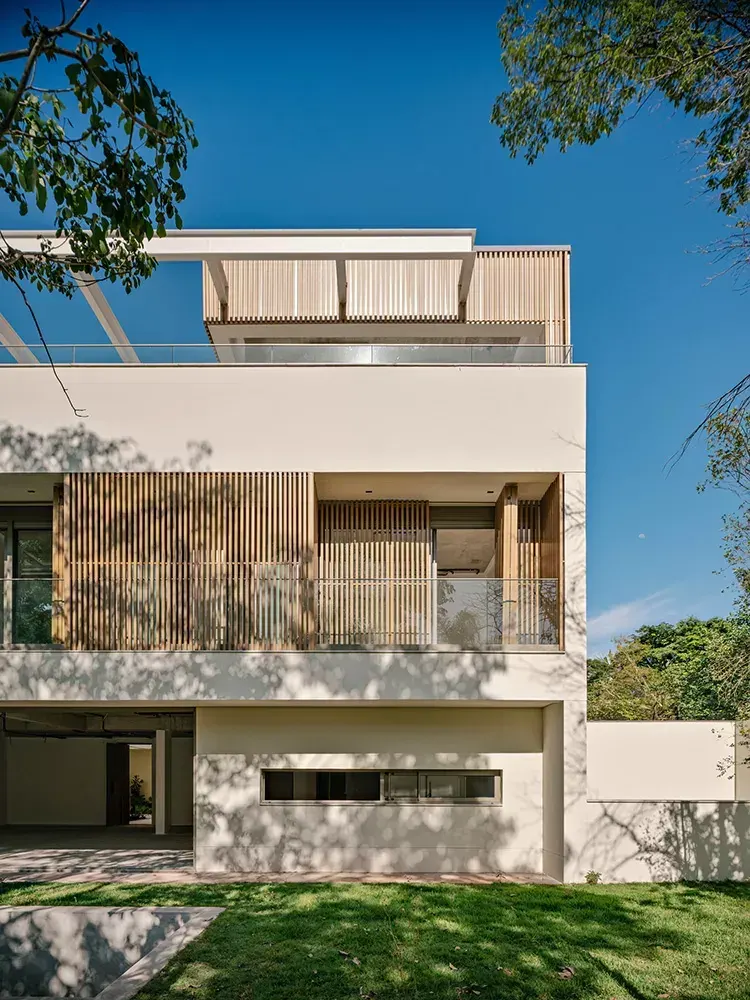 Uma casa do Kiná de frente, vista do jardim: o térreo aberto entre os pilotis, o volume branco das suítes com o ripado de madeira e o guarda-corpo de vidro e, no alto, a pérgula branca do rooftop contra o céu azul.