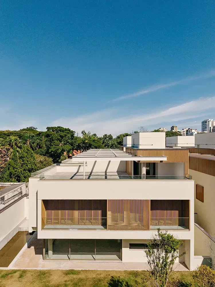 Uma das casas vista do alto: o volume branco com o ripado de madeira, o terraço do rooftop e, atrás, as árvores e os prédios do bairro sob o céu azul.