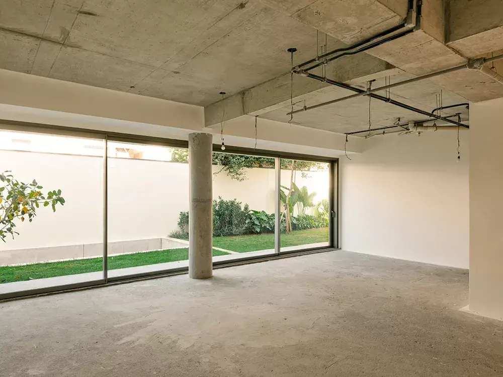 O térreo de uma casa por dentro, sem móveis: o teto de concreto com as instalações à vista, o piso cru e a parede de vidro aberta para o jardim.