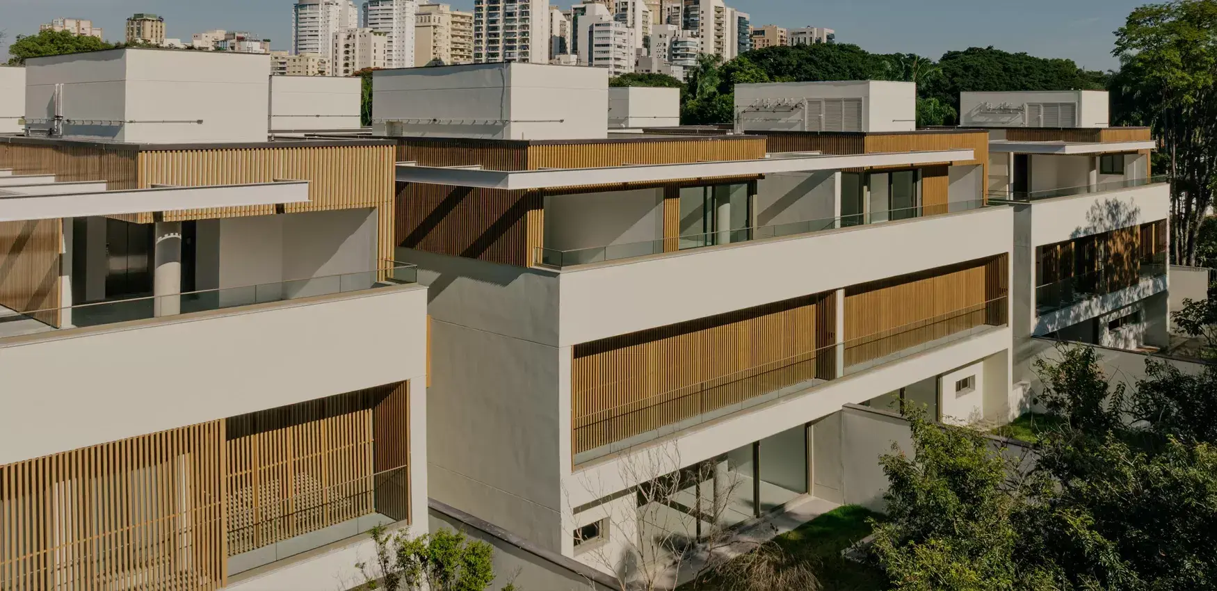 As casas do Kiná prontas, numa tarde de sol: os volumes brancos com o ripado de madeira e os guarda-corpos de vidro, entre as árvores do Alto de Pinheiros.