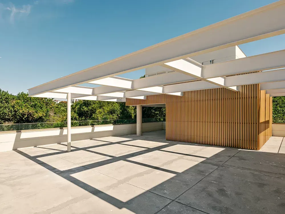 O rooftop de uma casa: a pérgula branca projetando sombras em grade sobre o piso claro, um volume de ripas de madeira e as copas das árvores além do guarda-corpo.