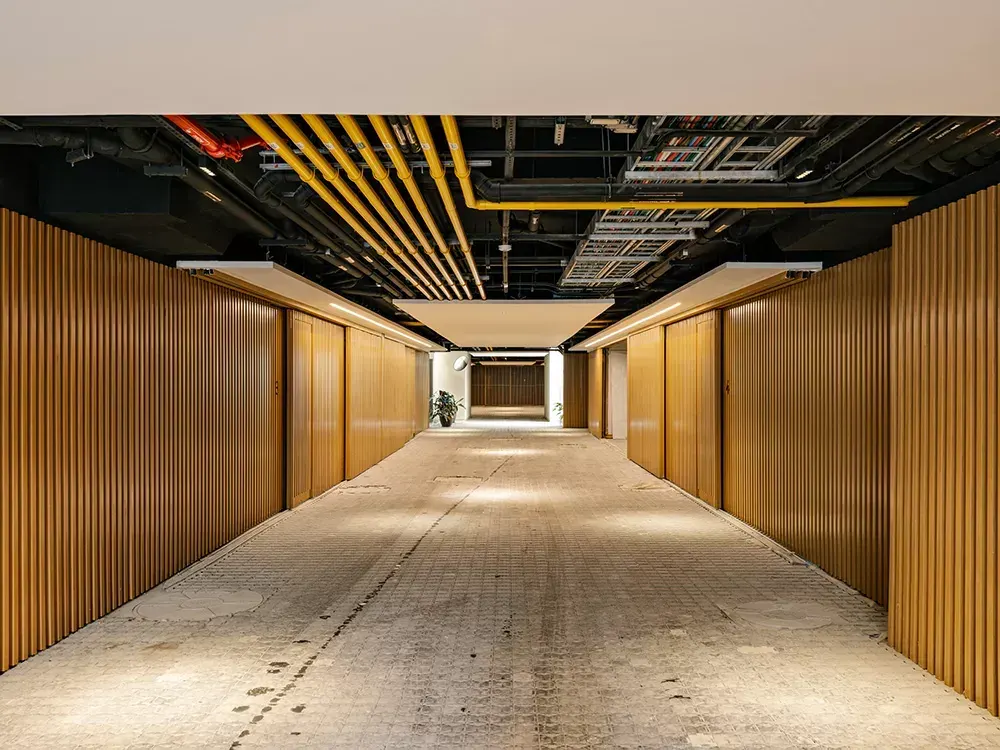 O corredor do subsolo do condomínio: o piso de concreto, as portas e os painéis de madeira dos dois lados e as instalações no teto.