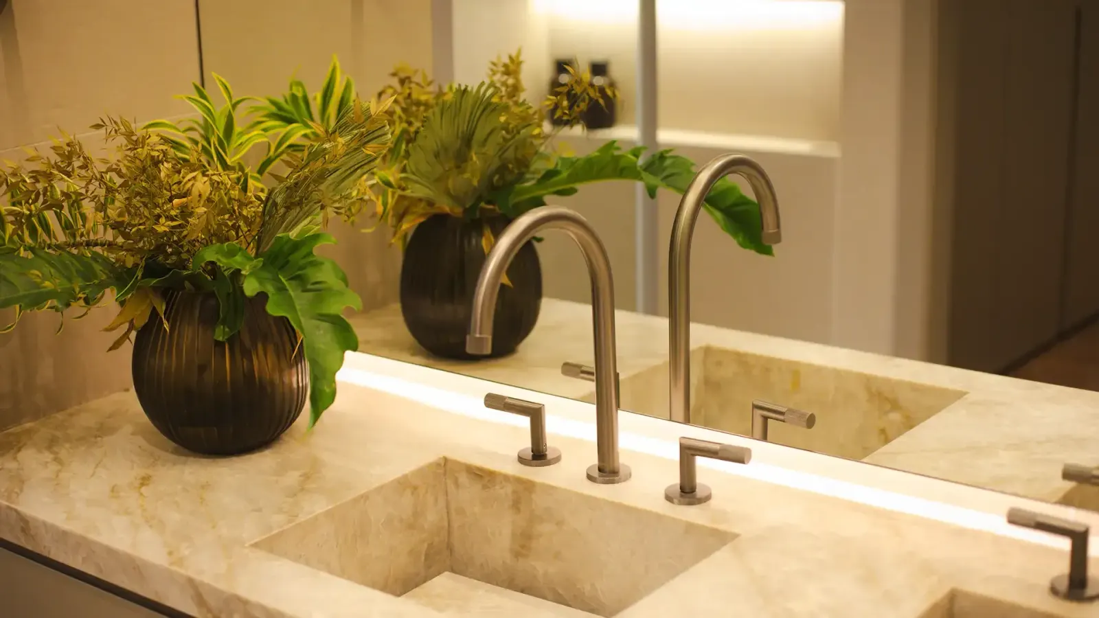 Um banheiro: a cuba esculpida na pedra clara, a torneira metálica e vasos de plantas na bancada.