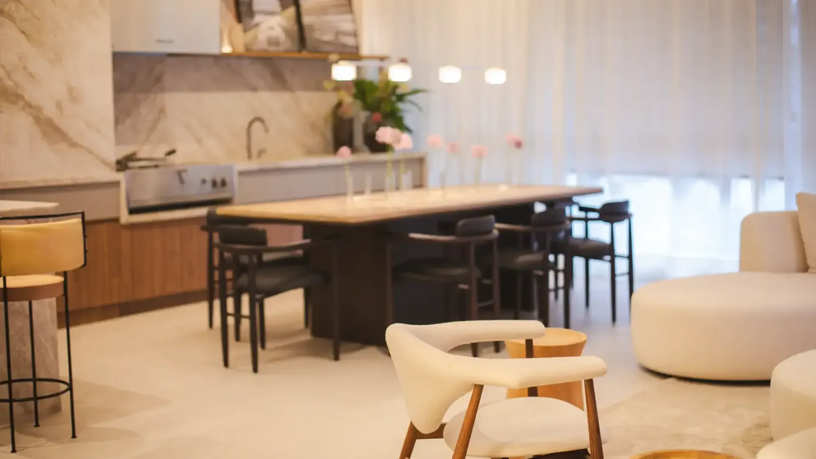 O estar e o jantar integrados: poltronas, o sofá curvo e a mesa longa, com a luz da janela pela cortina.