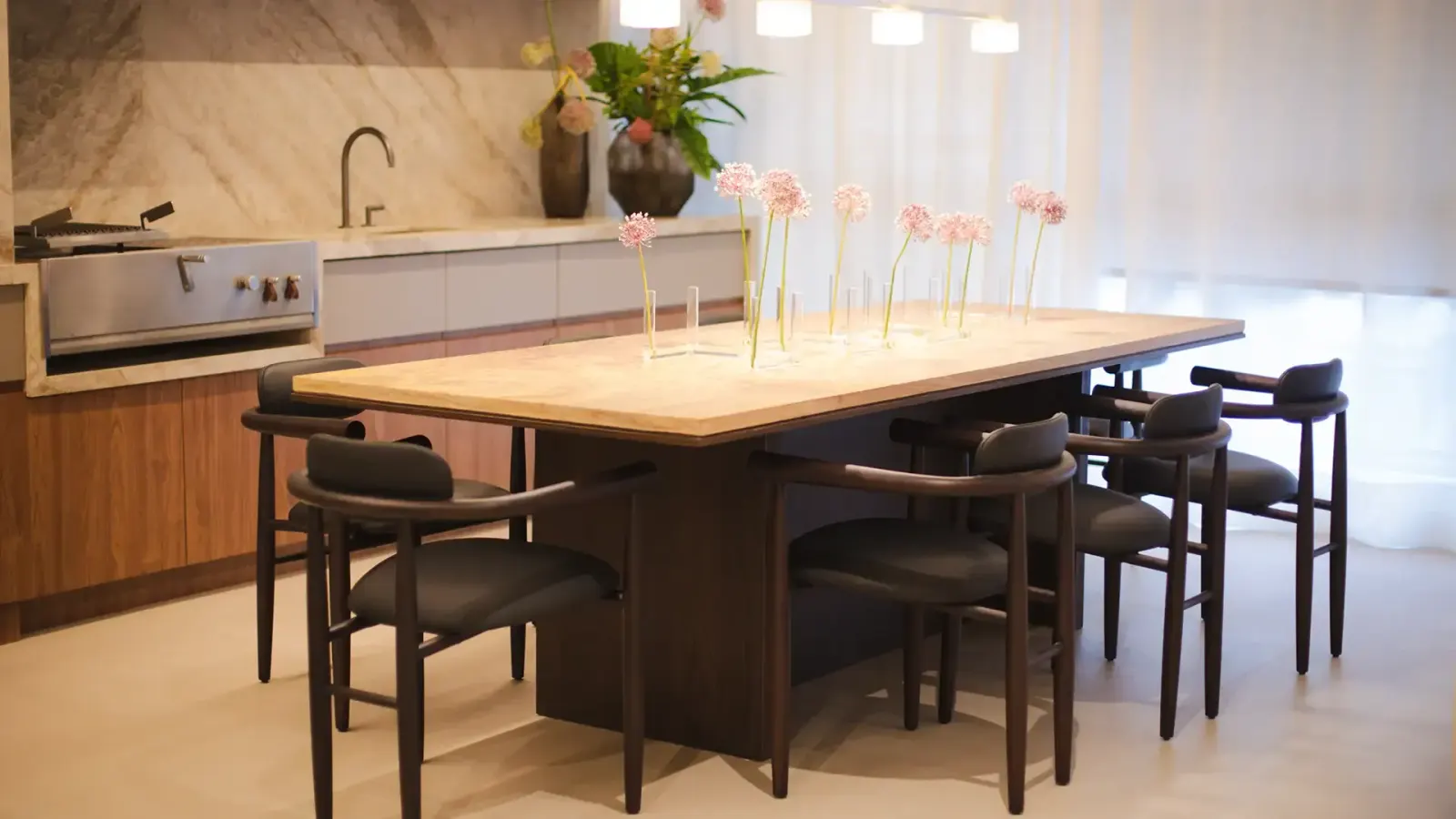 A mesa de jantar de pedra clara com cadeiras escuras, vasos finos de flores e a cozinha ao fundo.