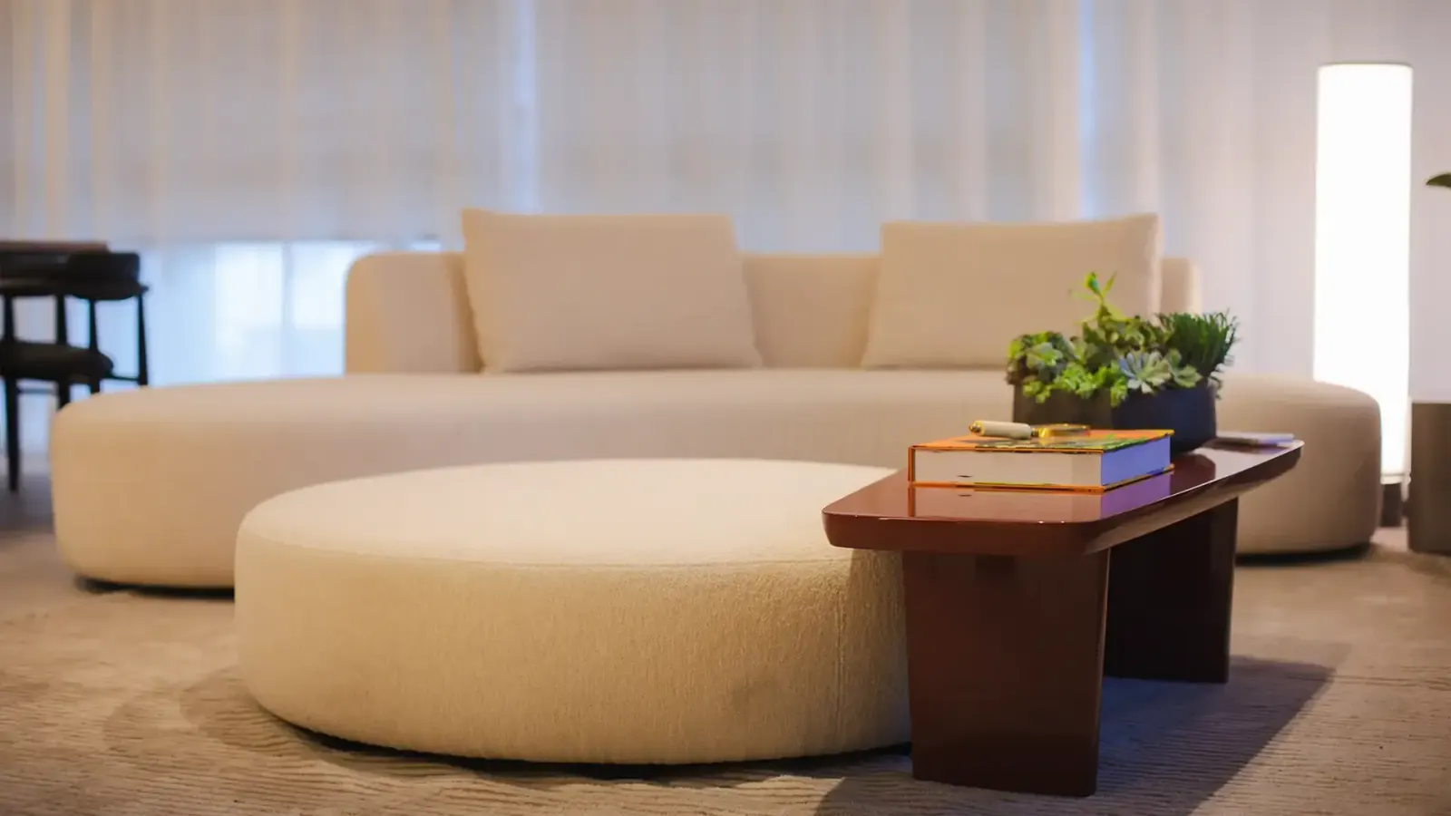 O living: um sofá curvo branco e um pufe redondo sobre o tapete claro, com a mesa de centro de madeira e a cortina ao fundo.