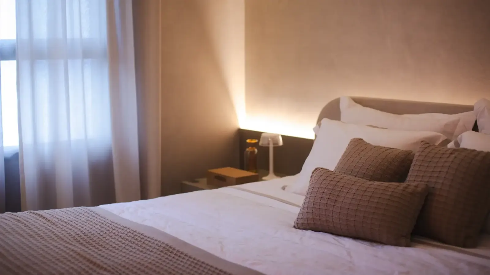 Uma suíte: a cama de lençóis brancos com almofadas, a luz baixa da cabeceira e a cortina clara.