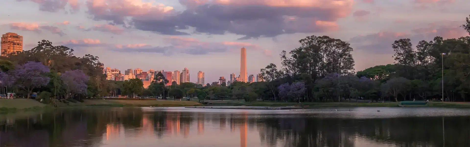 O lago do Parque Ibirapuera ao entardecer, com as árvores na margem e o obelisco e os prédios da cidade no horizonte.