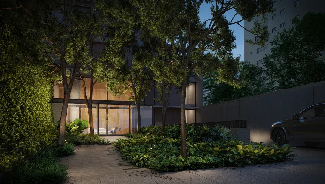 O jardim do embasamento à noite: árvores e folhagens iluminadas por baixo, um caminho de pedra e a base do prédio ao fundo.