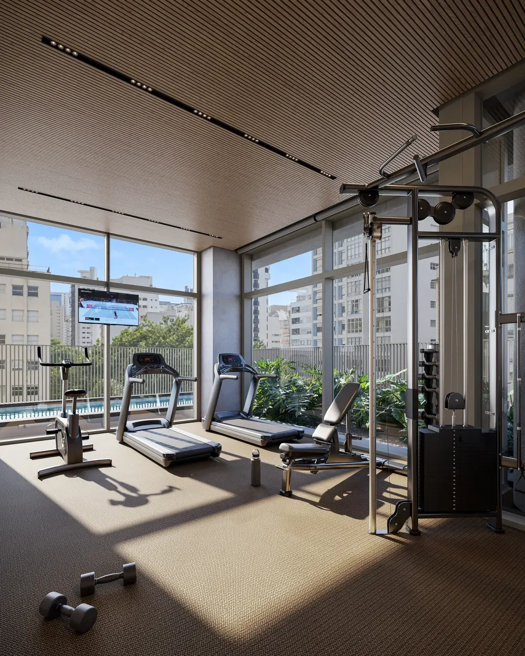 O fitness do residencial: a estação de musculação, a esteira e os halteres no chão claro, com a janela alta para o bairro.