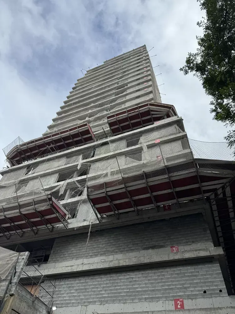A torre em obra vista da calçada, de baixo para cima: a estrutura de concreto inteira, as telas de proteção vermelhas nos andares de cima e as árvores da rua.