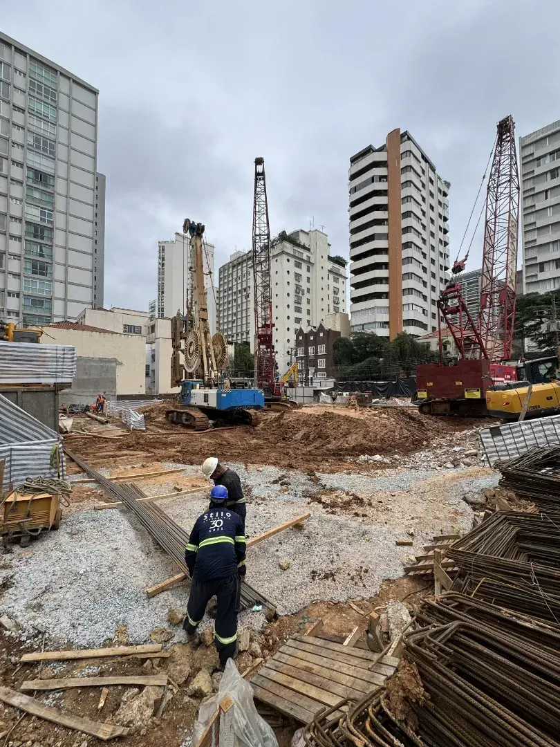 O terreno em obra: o chão de terra escavado, um operário de capacete e as máquinas de fundação, com os prédios vizinhos em volta.