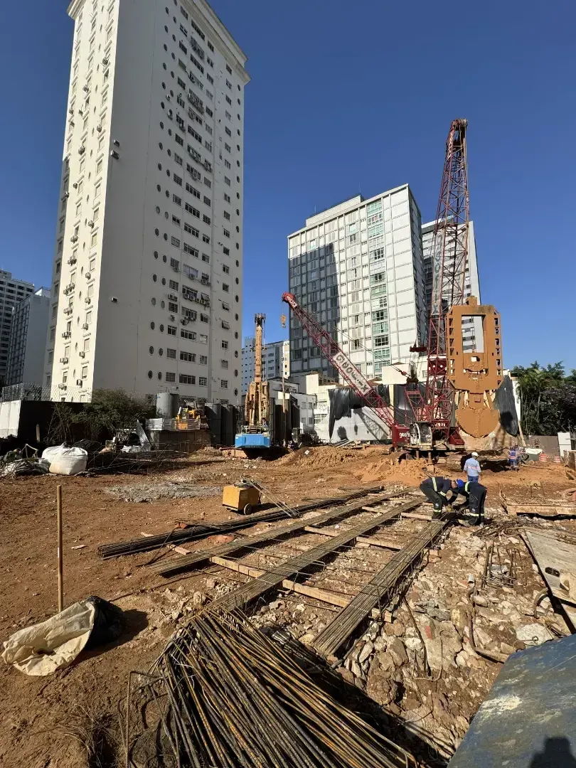O canteiro de dia, com o céu azul: as armaduras de aço no chão, a máquina de fundação e os prédios vizinhos.