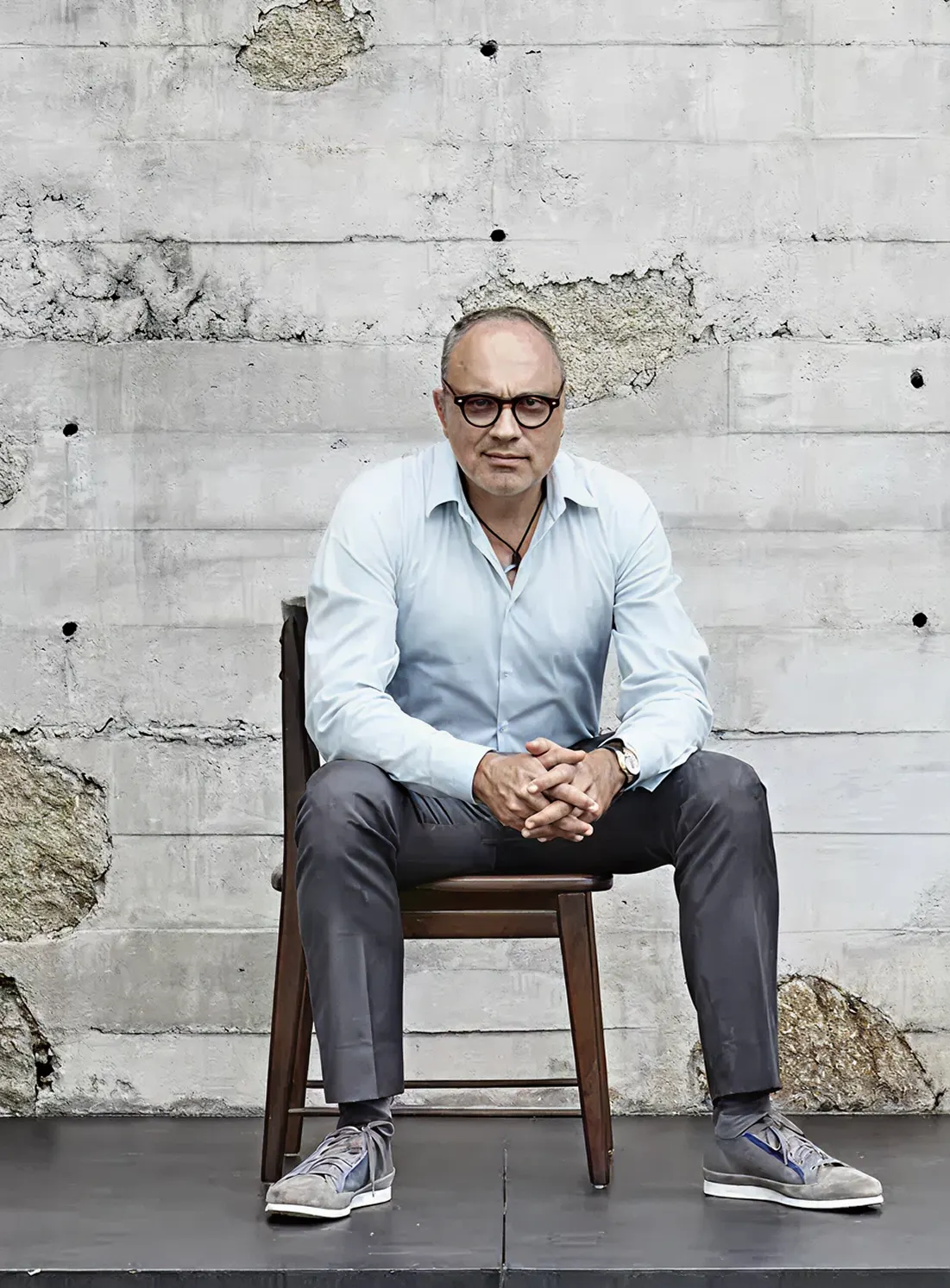 Arthur Casas sentado numa cadeira de madeira diante de um muro de concreto aparente, de camisa azul-clara e óculos de armação escura.