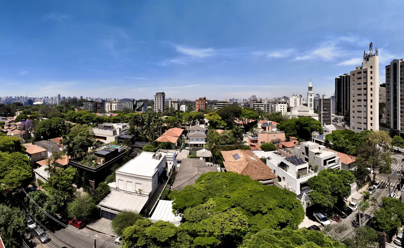 O bairro visto do alto: casas entre árvores, uma torre de igreja e, no horizonte, os prédios da cidade sob o céu azul.