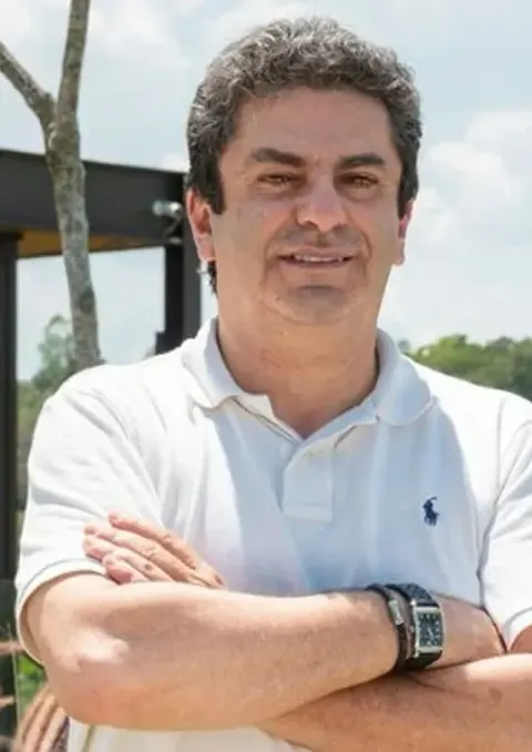 Roberto Riscala de camisa polo branca, braços cruzados, ao ar livre, com árvores ao fundo.