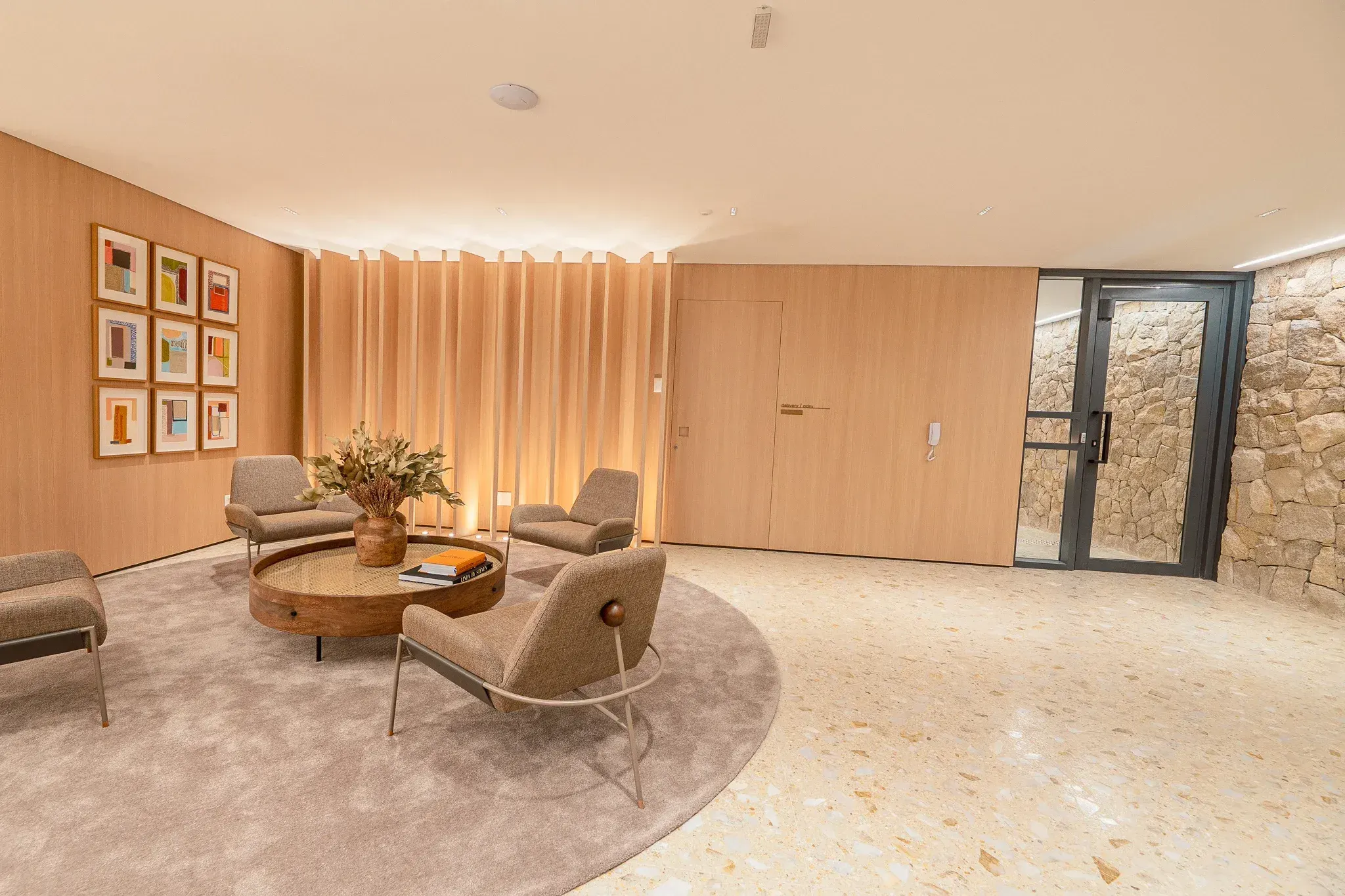 O hall social: os painéis de madeira com luz embutida, os quadros na parede, as poltronas claras em volta de uma mesa redonda sobre o tapete e o piso de granilite.