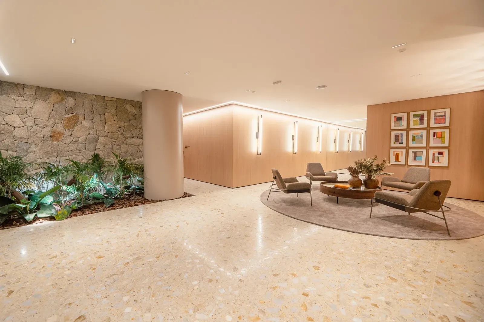 O hall social visto do outro lado: a parede de pedra com plantas embaixo, a mesa redonda com uma planta, as poltronas e a luz embutida no forro.