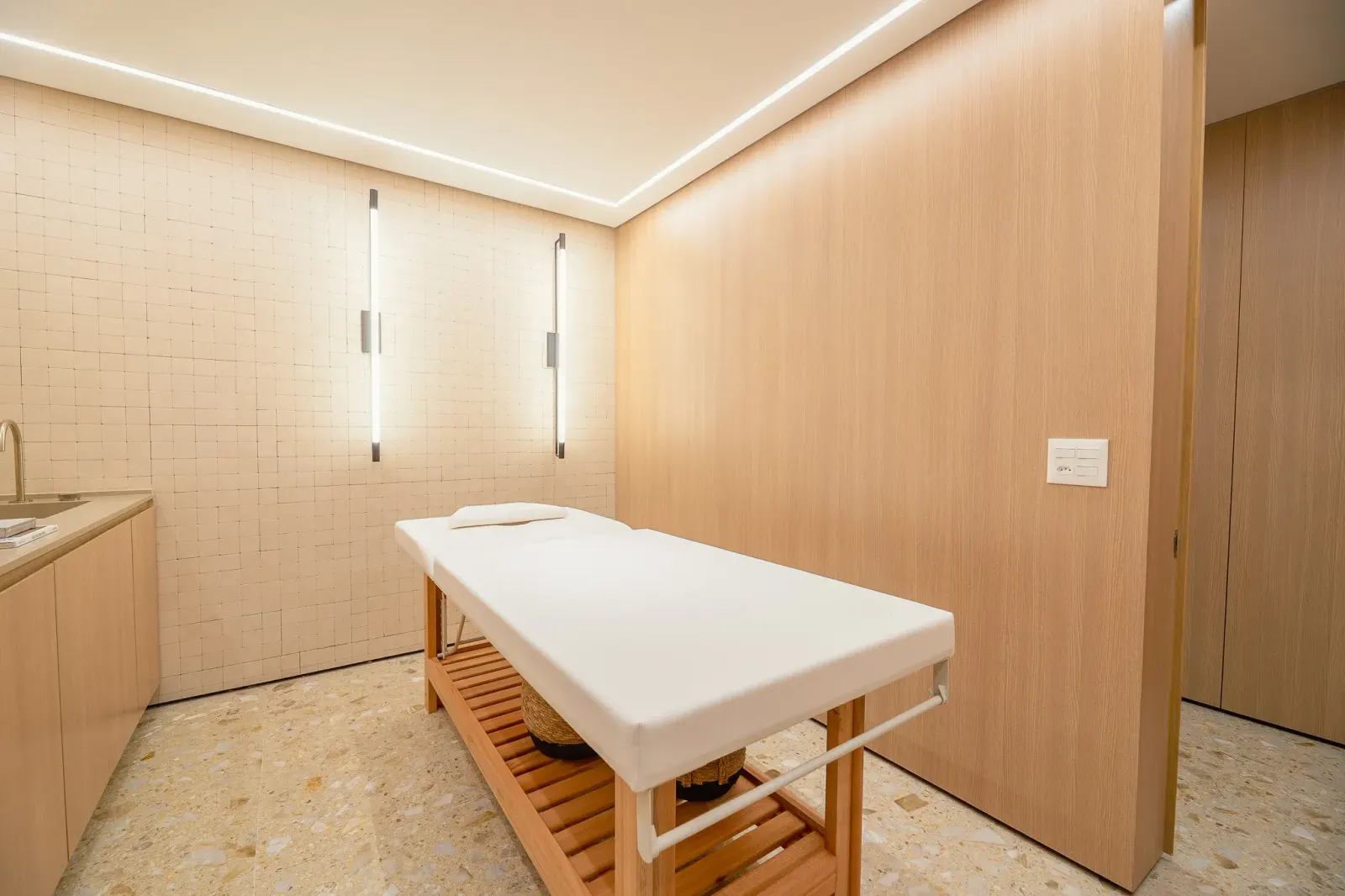 A sala de massagem: a maca branca, a parede de azulejo claro, os painéis de madeira, os quadros e um vaso com plumas secas.