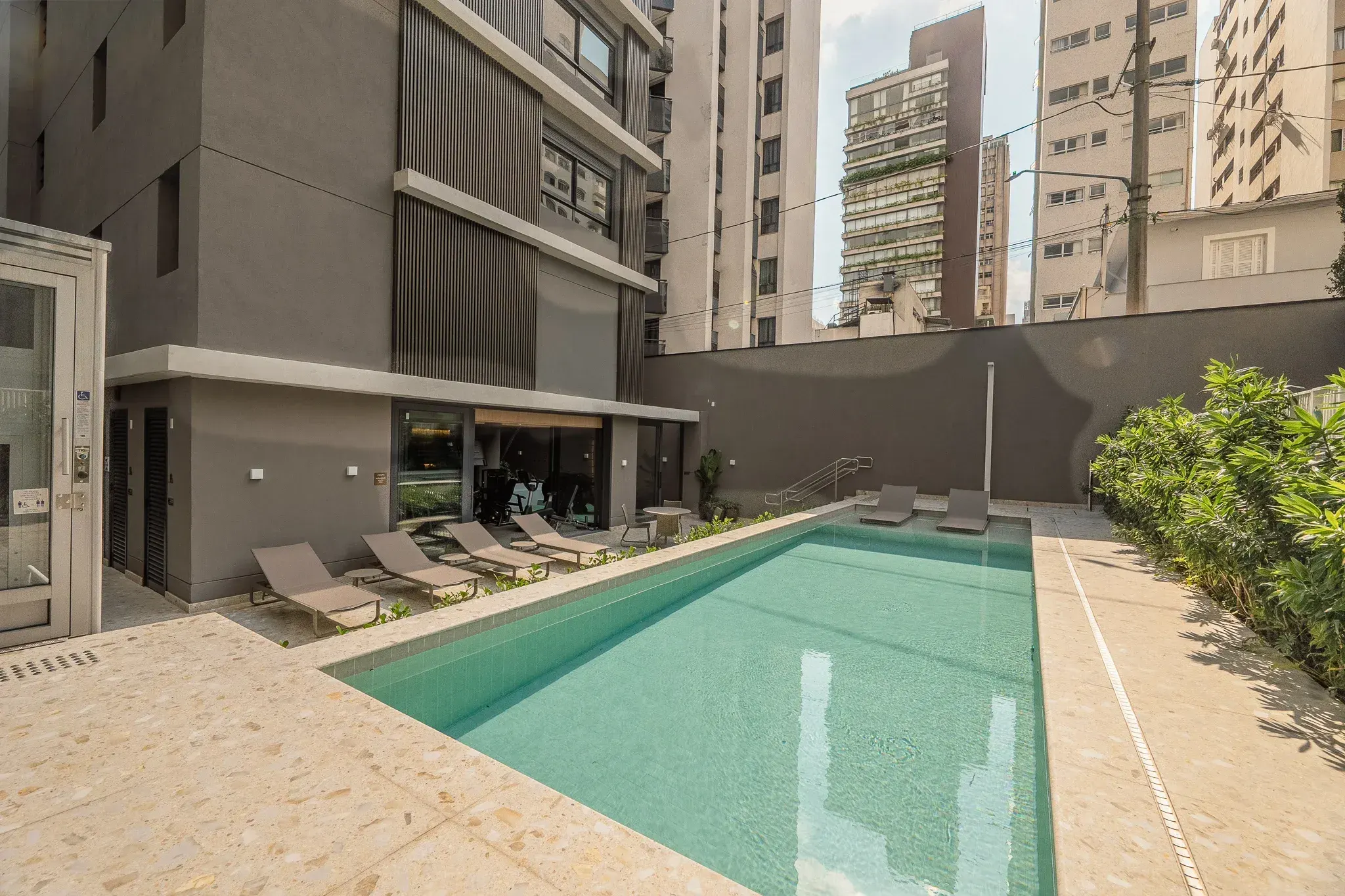 A piscina adulto no térreo, de água verde-clara, com o deque de pedra e as espreguiçadeiras de um lado, as plantas do outro e os prédios vizinhos em volta.
