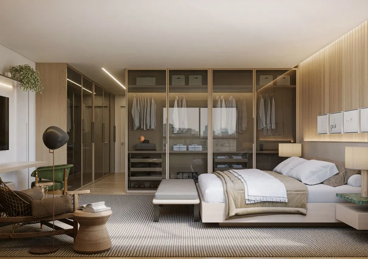A suíte master do apartamento: a cama com a cabeceira de madeira, o closet de vidro iluminado ao fundo e uma poltrona com mesa lateral ao lado.
