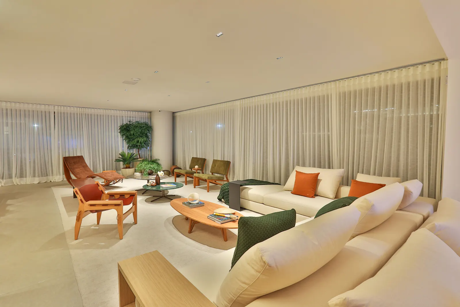 O estar do decorado com sofá claro em L, almofadas laranja e verdes, poltronas de madeira e a cortina leve na parede de vidro.