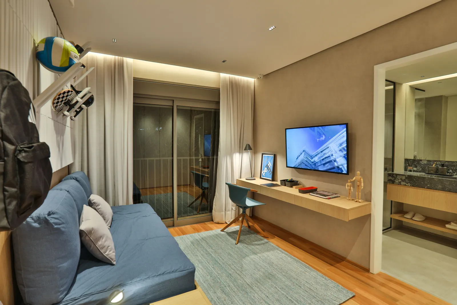 Um quarto do decorado montado como sala de jovem, com sofá-cama azul, escrivaninha, televisão e a porta de correr para a varanda.