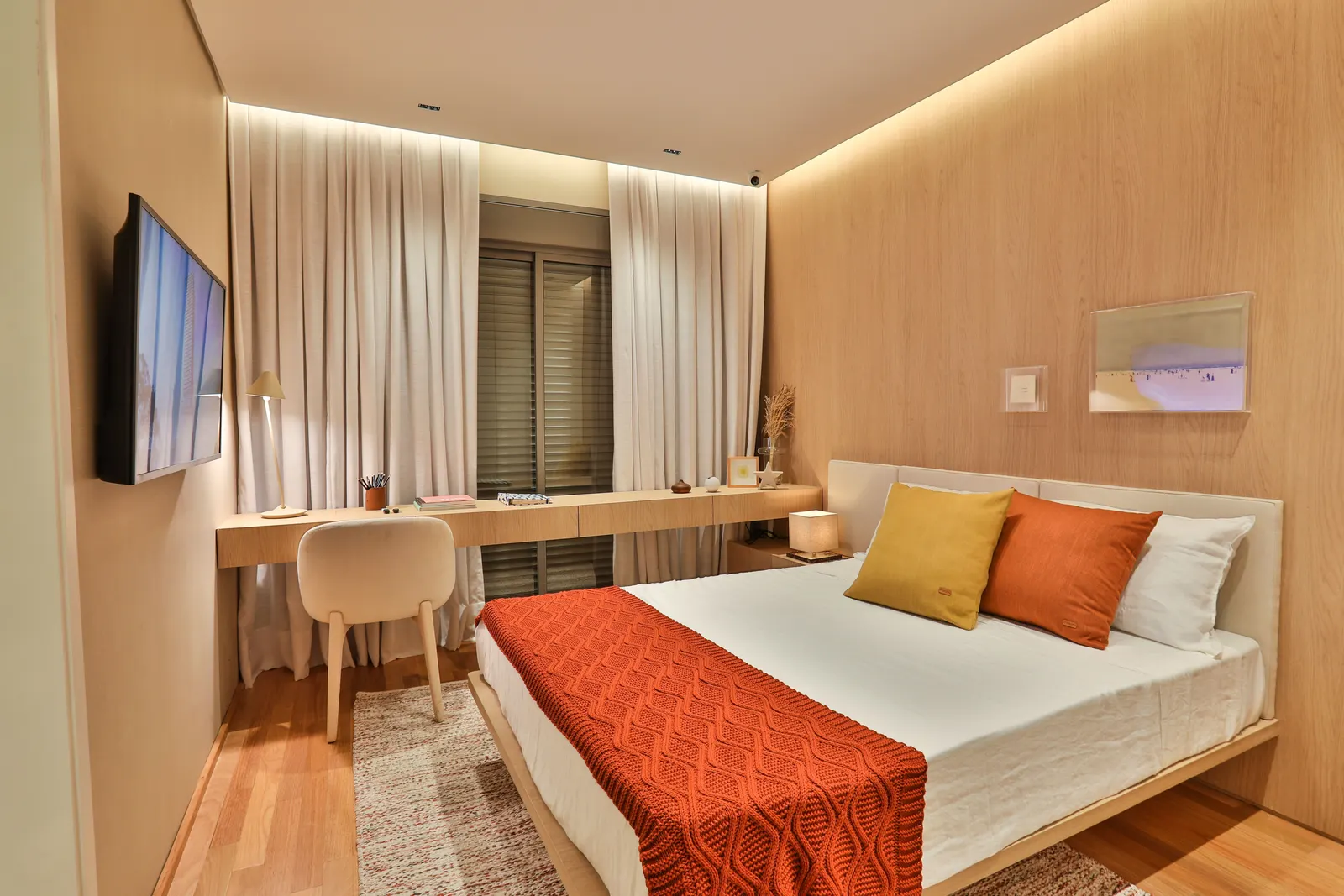 Uma suíte do decorado com cama de casal de manta laranja, painel de madeira iluminado, escrivaninha e a porta de correr com persiana.