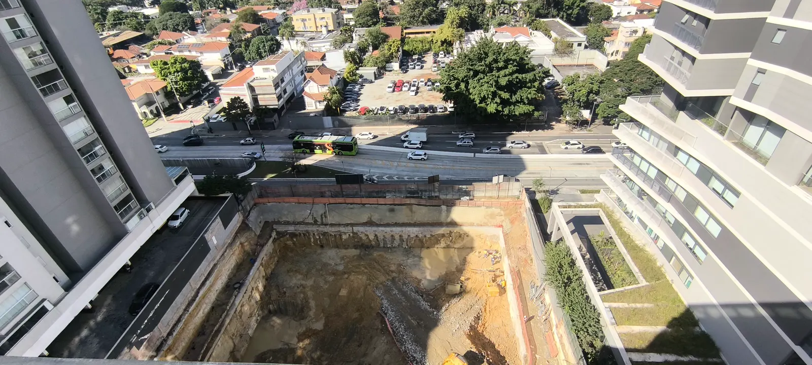 A obra vista do alto em junho de 2026: o terreno escavado com uma escavadeira amarela no fundo, a avenida em frente e os prédios vizinhos dos dois lados.