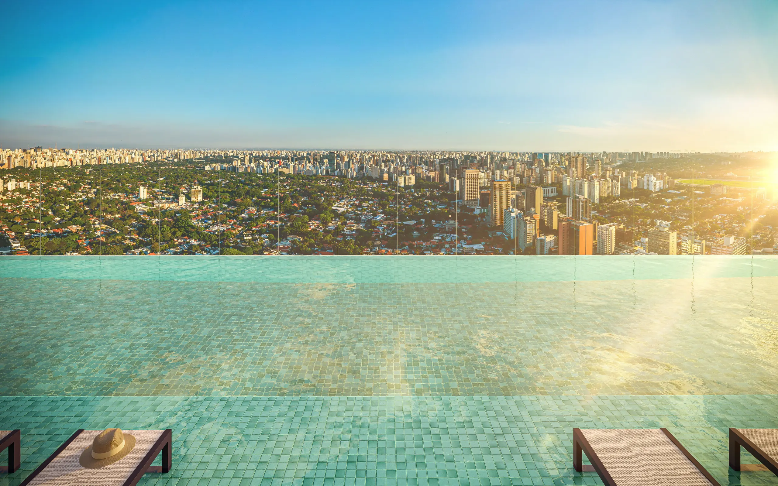 A piscina de borda infinita no rooftop, com a água verde-clara se encontrando com o verde dos Jardins e a cidade ao pôr do sol.