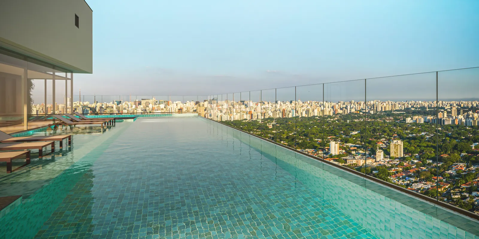 A piscina comprida ao longo do guarda-corpo de vidro, com espreguiçadeiras no deck e a cidade embaixo.
